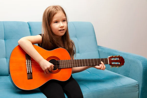 Young child, little school age girl playing classic acoustic guitar, practicing musical instrument sitting on the sofa Simple music education at home, child\'s hobbies and talents, portrait, interior.