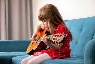 Küçük bir kız, genç bir okul çocuğu akustik gitar çalıyor, evdeki bir kanepede, iç mekanda, yan gözle müzik aleti çalıyor. Müzik eğitimi, hobi, çocukların yetenek kavramı.