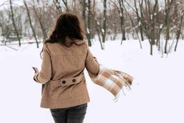 Elinde bej ceketli bir telefon olan kıvırcık saçlı, kahverengi saçlı bir kadın kışın sırtında duruyor. Yaşam tarzı, kız kaçıyor..