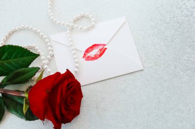 Kırmızı bir gül, üzerinde öpücük izi olan beyaz bir zarfın yanında yatıyor. Narin inciler yakınlarda sarılı. Aşkı ifade etmek için mükemmel, zarif ve romantik bir sahne yaratıyorlar..
