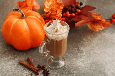 Sıcak bir içecek, üzerinde krem şanti ve baharatlar olan cam bardakta durur. Parlak turuncu bir balkabağı ve renkli sonbahar yapraklarının yakınında sıcak bir düşüş ortamı oluşur..