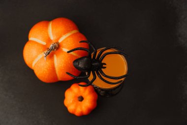 Turuncu balkabakları ürkütücü bir içecekle dolu bir bardağın yanında otururlar. Kenarına siyah plastik bir örümcek konur. Cadılar Bayramı temasını canlı renkler ve ürkütücü detaylarla güçlendirir..