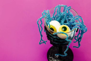 Parlak mavi şekerli iplikçiklerle dolu bir bardakta dinlenen iki şekergöz içeren ürkütücü bir tatlı. Dekor, canlı mor bir arka planla Cadılar Bayramı havası yaratıyor..