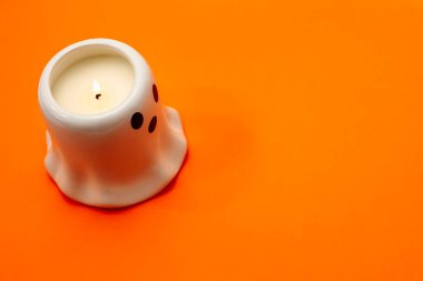 Canlı turuncu bir yüzey üzerinde siyah gözlü, küçük hayalet şeklinde bir mum duruyor. Bu büyüleyici dekoratif parça Cadılar Bayramı kutlamaları ya da temalı etkinlikler için mükemmel bir eğlence ve şenlik dokunuşu ekler..