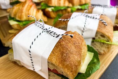 Delicious sandwiches are neatly wrapped with white paper and black twine. Each sandwich features fresh lettuce and tomato resting on a wooden board ready to be enjoyed for lunch.