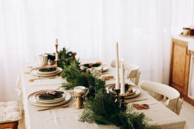 Christmas arranged dining table is set for a festive gathering. Greenery surrounds plates and candles creating a cozy atmosphere.