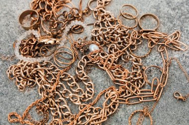 Çeşitli altın mücevher parçaları dokulu taş bir yüzeye yayılır. Koleksiyonda zincir halkalar ve inci bir kolye var. Hepsi birbirine dolanmış ve karmaşık bir düzenleme oluşturuyor..