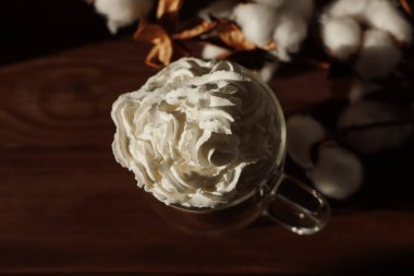 Krem şanti ile dolu temiz bir cam bardak ahşap bir yüzeye oturur. Yumuşak pamuk tokaları fincanın yanına dizilir sıcak ve davetkar bir atmosfer yaratır..