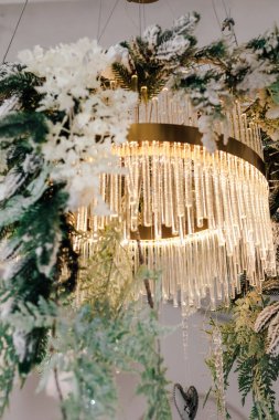 Tavandan sarkan göz kamaştırıcı bir avize. Kış yeşilliği ve beyaz çiçeklerle süslenmiş. Yumuşak ışıklandırma, bu güzel dekore edilmiş alanda mevsim için mükemmel bir ortam yaratır..
