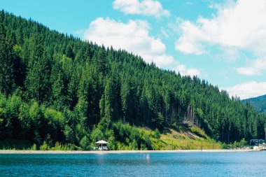 Sakin bir göl, parlak mavi gökyüzünü ve kabarık bulutları yansıtırken uzun yeşil ağaçlar kıyıya dizilir. Rıhtımda küçük ahşap bir pavyon duruyor. Huzurlu manzaraya cazibe katıyor..