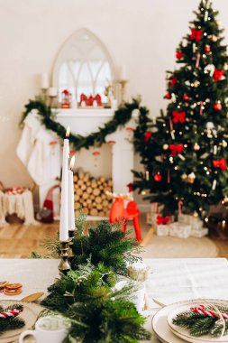 Güzelce düzenlenmiş bir masa şenlikli bir yemek için mumlar ve yeşillik içerir. Arka planda süslü bir Noel ağacı sıcak, neşeli bir atmosfer ekler oturma odasına..