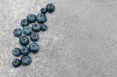 Bir grup taze yaban mersini desenli gri bir yüzeye saçılmıştır. Böğürtlenlerin koyu mavi rengi sağlıklı bir atıştırmalık ya da tatlı seçeneği sunan gri ile iyi uyumludur..