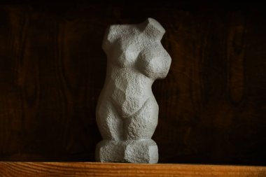 A unique sculpture of a female torso sits on a wooden shelf in a warm and inviting space. The artistic piece showcases a textured surface and graceful curves adding charm to its surroundings.