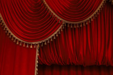 Deep red velvet curtains hang elegantly with intricate golden tassels creating a dramatic backdrop for a theater performance. The luxurious fabric adds to the artistic atmosphere of the stage.