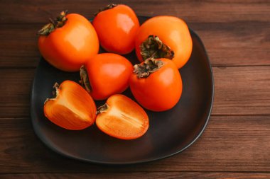 Bright orange persimmons sit on a black plate adding a vibrant touch to the kitchen. Some fruits are whole while one is sliced to reveal its juicy interior.