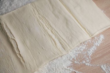 Soft layers of pastry dough are spread out on a wooden countertop sprinkled with flour. The kitchen is bright and cozy perfect for baking delicious treats.