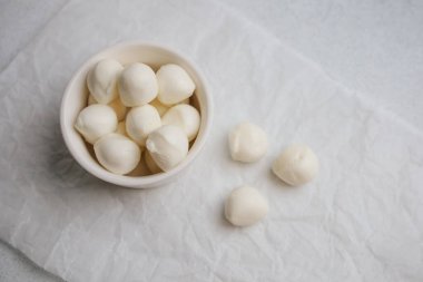 Small creamy mozzarella balls are neatly arranged in a white bowl on a light surface. Several balls are scattered around the bowl creating an inviting display of fresh cheese.