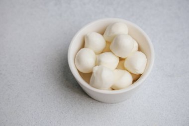 Soft mozzarella balls are neatly placed in a small white bowl showcasing their creamy texture. The bowl is set on a light surface making the cheese look appetizing and fresh.
