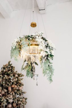 A beautifully designed chandelier hangs from the ceiling decorated with flowers and greenery. Below a festive Christmas tree is adorned with shiny ornaments creating a cheerful holiday atmosphere.