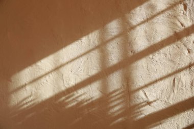 Sıcak güneş ışığı boşluklardan süzülerek dokulu bir duvara uzatılmış gölgeler bırakır. Nazik ışık yüzeyin ince detaylarını güçlendirir. Odada davetkar bir atmosfer yaratır..