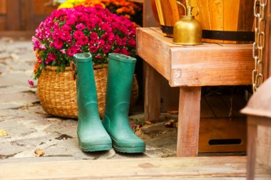 Ahşap bir bankın yanında yerde duran yeşil lastik botlar. Örülmüş sepetlerdeki renkli çiçekler bahçeye cazibe katar. Güneş ışığı, canlı bir açık hava manzarasını gözler önüne seriyor..