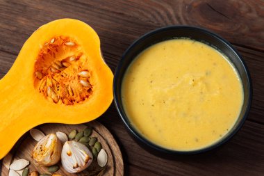 Bir tereyağlı balkabağı, bir kase kremalı çorbanın yanındaki tohumları göstererek açılır. Malzemeler ahşap bir yüzeyde sergileniyor. Mutfak dekoru yemek atmosferine katkı yapıyor..