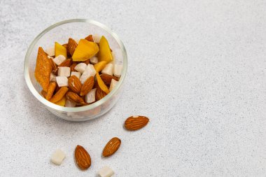 Bir kase fındık ve kurutulmuş meyve karışımıyla dolu hafif bir yüzeyde oturur. Kurutulmuş mango parçaları ve yumuşak şeker küpleri renkli bir atıştırmalık sunar..