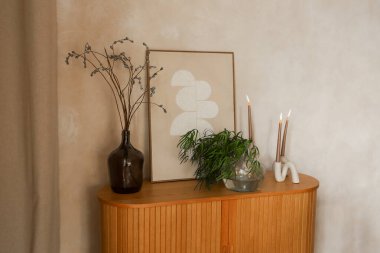 Ahşap bir dolapta çerçevelenmiş bir sanat eseri ve bitkilerle dolu cam bir vazo var. Tutucularda iki mum var. Yumuşak ışık odayı doldurur ve basit düzenlemeleri güçlendirir..