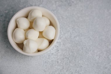 Beyaz bir kase düzgünce düzenlenmiş mozzarella peyniri küçük topları içerir. Kase açık renkli bir yüzeye dayanır. Yemeklerin hazırlandığı ya da servis edildiği bir mutfak ya da yemek alanı öneriliyor..