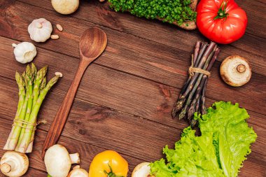 Kırsal bir ahşap masada ahşap bir kaşığın etrafında çeşitli taze sebzeler düzenlenir. Sahnede sebze, domates, mantar ve kuşkonmaz karışımı yemek hazırlığı öneriliyor..