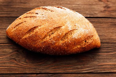 Bir somun kahverengi ekmek mutfaktaki ahşap bir masanın üzerinde duruyor. Ekmeğin kabuklu bir yüzeyi ve görünür kesikleri var..