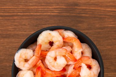 Siyah bir kase düzgünce düzenlenmiş pişmiş karides içerir. Karideslerin yemeye hazır olduklarını gösteren pembe bir tonu var. Kase ahşap bir masaya konur ve yemek için basit bir ortam sunulur..