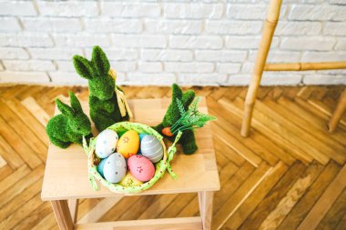 Tahta bir masanın üzerinde renkli yumurtalarla dolu bir sepetin yanında üç yeşil tavşan figürü duruyor. Dekor, ahşap döşemeli ve açık renkli duvarlı parlak bir oda içeriyor..