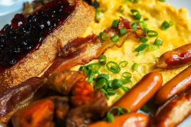 Kahvaltı tabağında yeşil soğanlı çırpılmış yumurta kıtır pastırma sosisi ve çilek reçelli bir dilim tost var. Yemekler beyaz bir tabakta servis edilir ve basit bir yemek oluşturur..