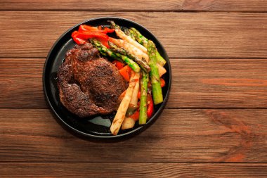 Büyük bir biftek, kuşkonmaz ve dolmalık biber de dahil olmak üzere çeşitli ızgara sebzelerin yanında siyah bir tabakta oturur. Ayarlar, sıradan bir yemek sahnesini gösteren ahşap bir masa..