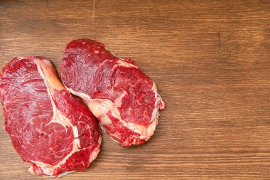 Ahşap bir yüzeye iki çiğ et kesiği yerleştirilir. Sahne pişmeye hazır taze etleri gösteriyor. Sığır etine odaklı basit bir mutfak düzeneği gibi görünüyor..