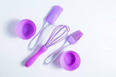 Beyaz bir yüzeyde mor pişirme araçlarının bir koleksiyonu sergilenmektedir. Set bir spatula çırpma fırçası, bir fırça ve silikon kaplar içeriyor..