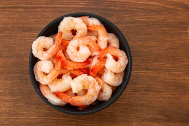 Büyük karidesler, ahşap bir yüzeyde, siyah bir kasede düzenlenir. Karides taze ve servis için hazır görünüyor. Bu düzen evde bir yemek ya da atıştırmalık için uygundur..