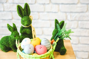 Çimenden yapılmış üç yeşil tavşan parlak boyalı yumurtalarla dolu bir sepetin yanında otururlar. Ayar, neşeli bir bahar kutlaması öneren hafif arka planlı basit bir masayı gösteriyor..