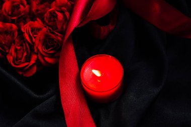 Bir demet kırmızı gül, siyah bir kumaş yüzeyinde kırmızı bir mumla yan yana oturur. Kırmızı bir kurdele güllerin etrafına dolanır ve akşam için uygun romantik ve sıcak bir atmosfer yaratır..