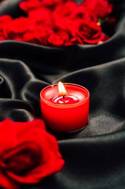 Kırmızı bir mum, koyu bir kumaş üzerinde taze kırmızı güllerle nazikçe yanar. Sahne akşam çekilir titreyen ışık ve canlı renklerle ılık bir atmosfer yaratır..