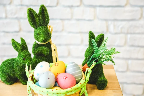 Çimenden yapılmış üç yeşil tavşan parlak boyalı yumurtalarla dolu bir sepetin yanında otururlar. Ayar, neşeli bir bahar kutlaması öneren hafif arka planlı basit bir masayı gösteriyor..