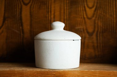 Yuvarlak beyaz bir kap ahşap bir rafta duruyor. Kutunun basit bir kapağı ve pürüzsüz bir yüzeyi var. Arkaplanda, uzayda sıcak ve davetkar bir atmosfer yaratan ahşap paneller yer alıyor..