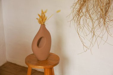 Kahverengi bir vazo kurumuş çiçekleri tutar ve basit bir odadaki ahşap bir tabureye oturur. Arka plan, vazolara ve içindeki çiçeklere odak veren düz bir duvar..