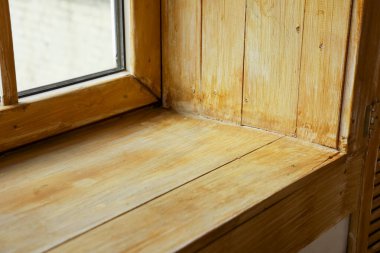 Tahta pencere pervazına yakın bir görüntü, doğal tahıl ve dokuyu gösteriyor. Pencere açık, odanın ışıkla dolmasına izin veriyor. Sıcak ahşap, etrafındaki duvarla çelişiyor..