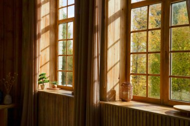 Güneş ışığı, ahşap bir odadaki büyük pencerelerden süzülür. Yeşil ağaçlar ve açık hava manzarası görülebilir. İçerideki bitkiler dekoratif eşyalar ve perdeler basit bir atmosfer yaratır..