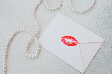 Açık bir yüzeyde beyaz bir zarf var. Önünde kırmızı bir dudak izi var. Beyaz inci (ler) dizili olduğu zaman, Bu sahne harflerle veya notlarla ilgili basit ve eğlenceli bir anı yakalar..