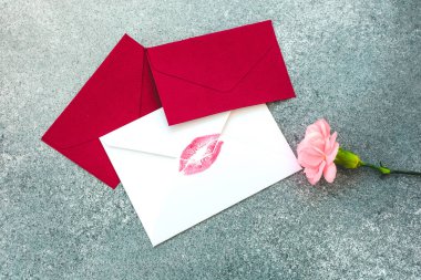 Üzerinde öpücük izi olan beyaz bir kartın yanında iki kırmızı zarf var. Gri bir yüzeyde pembe bir çiçek onların yanında duruyor..