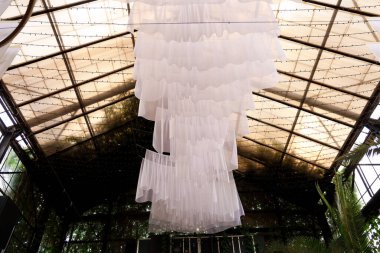 Kapalı bir alanda tavandan sarkan beyaz kumaş eşsiz bir tasarım yaratıyor. Yapının, doğal ışığın boşluğu doldurmasına olanak sağlayan açık bir çatısı var. Bitkiler görülebilir..