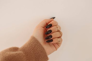 Bir el, uzun siyah tırnaklar ve parmaklarında çoklu altın yüzüklerle sergilenir. El, hafif ve nötr bir arkaplanda konumlandırılmış. Tırnakları göstermek için parmaklar hafifçe kıvrılır..